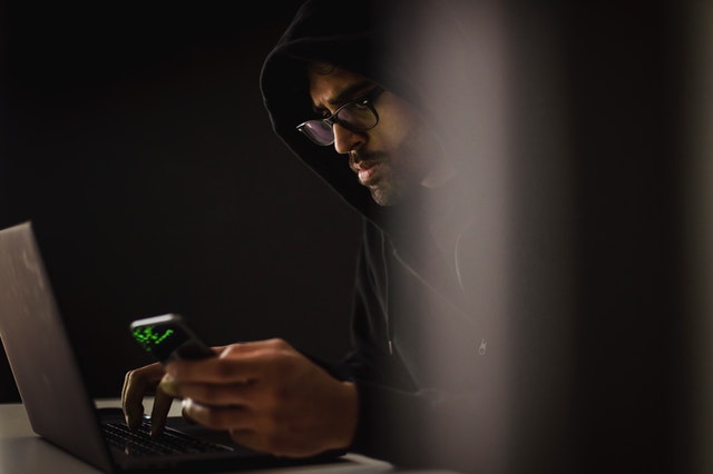crop-ethnic-hacker-with-smartphone-typing-on-laptop-in-darkness